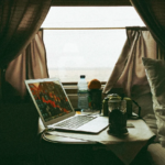 Un escritorio improvisado junto a una ventana con cortinas, donde hay una laptop encendida, una taza de café y una cámara fotográfica sobre la mesa.