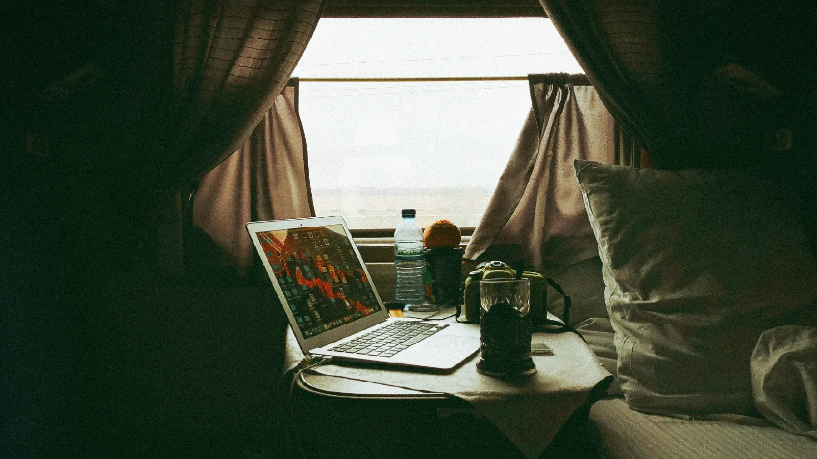 Un escritorio improvisado junto a una ventana con cortinas, donde hay una laptop encendida, una taza de café y una cámara fotográfica sobre la mesa.